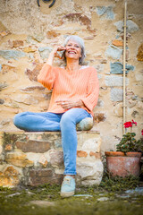 Senior woman laughing on the phone while sitting outside against a stone wall