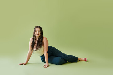 A beautiful plus size woman poses confidently in a corset and stylish pants against a rich green background.