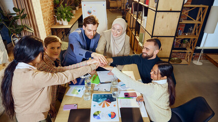 Group of multicultural colleagues join hands in team huddle around desk, symbolizing unity and...