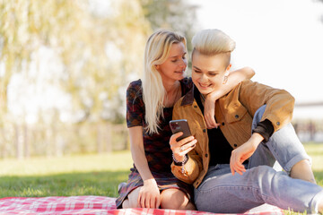 beautiful young lesbian couple in summer on the grass checkered blanket sitting hugging watching social networks relaxation of a couple in love