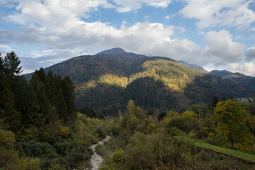 vista panoramica da fondovalle su un ambiente di montagna nell'Italia settentrionale, con alte montagne verdi, coperte da immensi boschi di conifere, nella penombra, di pomeriggio, con cielo nuvoloso