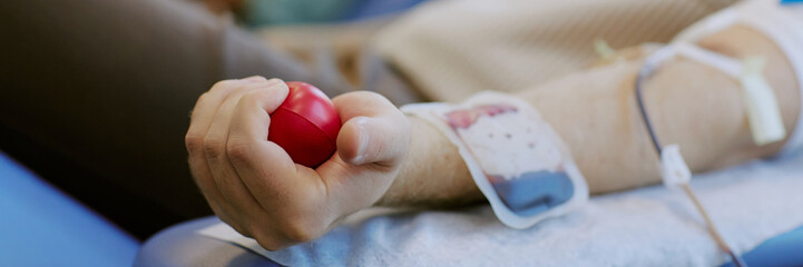 Person's hand squeezing red stress ball while donating blood, iv tube inserted in arm. Image suggests process of blood donation, emphasizes healthcare and community service