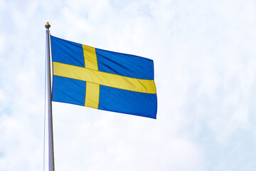 Swedish flag waving against a bright sky. The national flag of Sweden, featuring a golden cross on a blue background,  flying on a flagpole under a partly cloudy sky. Copy space