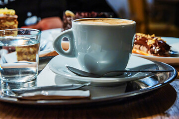 Fototapeta premium A closeup of a ceramic coffee cup on a silver tray with an eclair and a glass of water on the side. A cozy café setting with cappuccino with pastries.