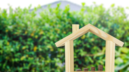 Fototapeta premium A wooden house model with coins arranged in ascending order inside, set against a blurred background of bushes, symbolizing financial growth and investment for home ownership.