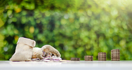 Brown burlap sacks filled with coins arranged in ascending order and some scattered on a white surface, with a blurred background of bushes, representing growth in savings and financial planning.