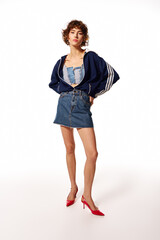 Fototapeta premium A young woman showcases her stylish denim attire and vibrant red heels with a confident expression.