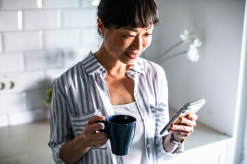 Woman smiling while checking her phone and holding a coffee mug in a bright kitchen