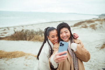 Two diverse female friends taking a selfie on a beach during winter