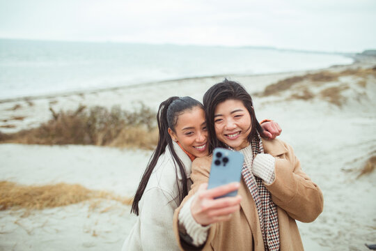 Two diverse female friends taking a selfie on a beach during winter - Powered by Adobe