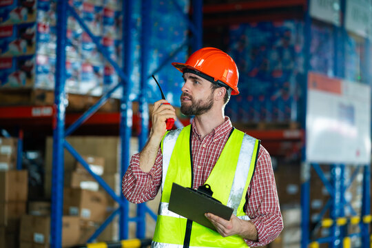 A man in a yellow vest and orange hat is standing in a warehouse. He is holding a clipboard and a walkie talkie - Powered by Adobe