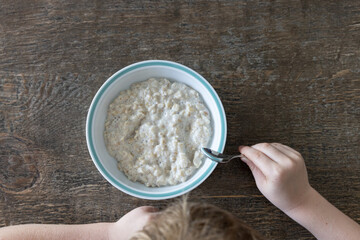 Freshly cooked oatmeal in a white porcelain bowl with blue rim on a rustic wooden dining table. A young light haired boy is sitting at the table holding a spoon about to start eating.