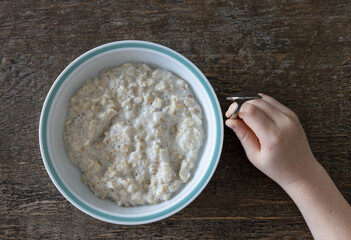 Freshly cooked oatmeal in a white porcelain bowl with blue rim on a rustic wooden dining table. Child's hand holding a small spoon about to start eating.