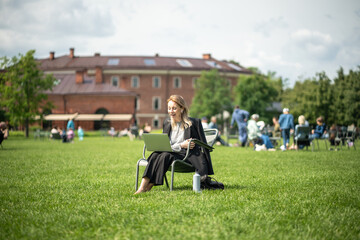 Fototapeta premium Young woman student or businesswoman sits on lounge chair in summer park with laptop and having video conference. Online webinar, video call, communication with laptop, outdoor business meeting. 