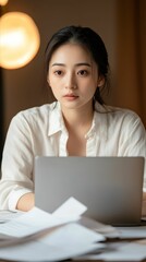 A young Asian businesswoman working late at her desk, focused on her laptop with papers and reports spread around, symbolizing dedication