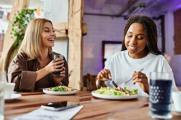 A smiling woman with braided hair enjoys a meal while her girlfriend looks on with delight at a cafe.