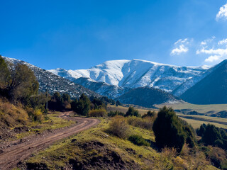 A dirt road winding through the mountain slopes in Kyrgyzstan, with snow-capped peaks in the background and a clear blue sky.