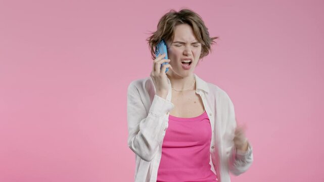 Annoyed young woman quarreling while talking on mobile phone. Angry lady in stress. Pink studio background. Conflict, dispute, negative.