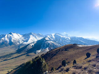 Panoramic view of snow-covered mountain ranges in Kyrgyzstan under a clear blue sky, with green valleys and autumn forests at the foothills