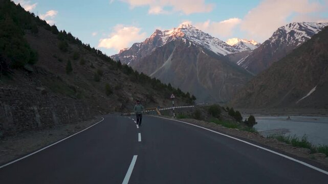 Slow motion shot of an Indian male tourist running towards the camera on a road in front of the snowy Himalayas during the sunset at Jispa in Lahaul and Spiti district, Himachal Pradesh, India.