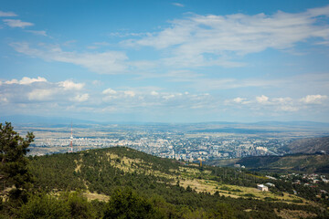 Clear mountain view of Tbilisi, Georgia, with rolling hills in the foreground and a partly cloudy sky above. The city stretches into the distance, framed by a scenic horizon