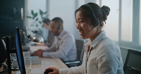 Call center office: Portrait of friendly Asian female hotline operator talking on headset, providing online customer assistance, looking at camera. Multiethnic team of technical support specialists.