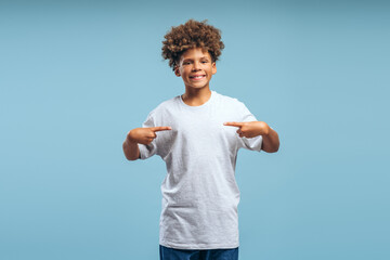 Portrait of smiling, positive African American boy wearing stylish white t shirt, pointing on mockup