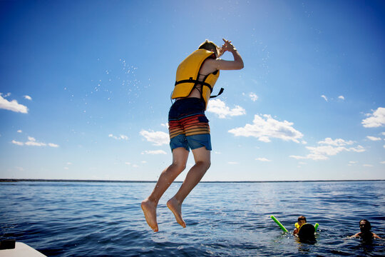 Young boy jumping off a boat and swimming in a lake with a lifejacket with his father and brother watching from the water, Lac Ste. Anne; Alberta Beach, Alberta, Canada
