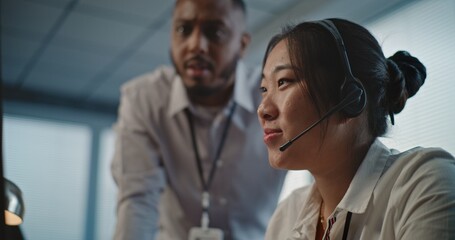 Call center office: African American technical support specialist helps Asian colleague in headset....
