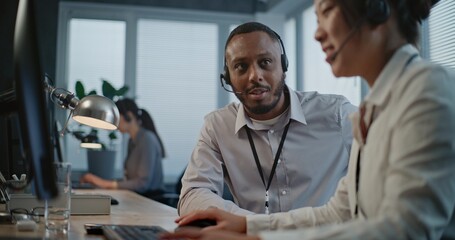 Call center office: Two multiethnic hotline operators in headsets discuss work, use computer having conversation with client on call. Technical support specialists working in online customer support.