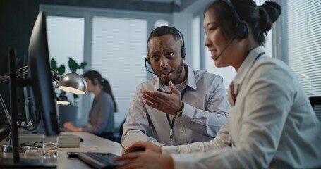 Obraz premium Call center office: Two multiethnic hotline operators in headsets discuss work, use computer having conversation with client on call. Technical support specialists working in online customer support.