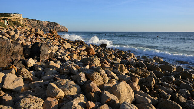 Surf splashes up on the rocks on Praia do Zavial along the coast of the Algarve region of Portugal, with swimmers enjoying the North Atlantic ocean in the background; Raposeira, Faro, Portugal