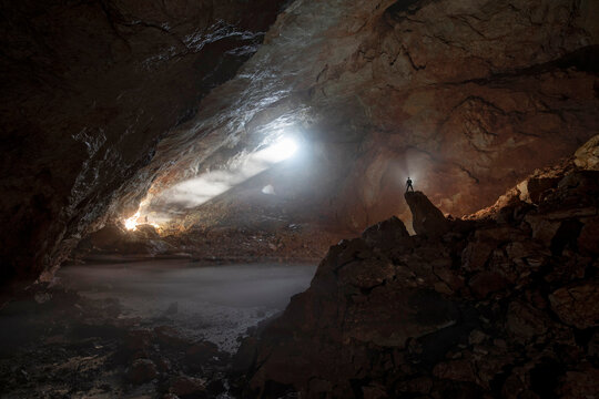 Scientist stands in silhouette on a rock formation inside an ice cave of the European Alps, viewing the natural beauty that is vulnerable to climate change; Asiago, Veneto, Austria