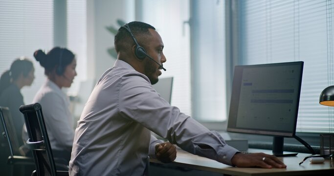 Modern call center office: African American consultant sits down at computer, puts on headset, uses computer. Team of diverse technical support operators talking providing online help for customers.