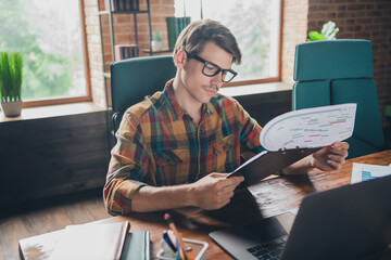 Photo of handsome young man wear checkered shirt sit table workplace read stats indoors office loft...