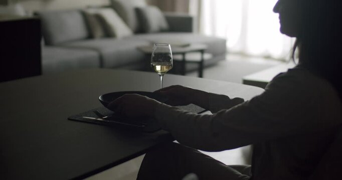 A serene woman sitting alone at a dining table with a glass of wine in a cozy living room, reflecting mood and solitude. Serene Woman Dining Alone with Wine Glass in Cozy Living Room