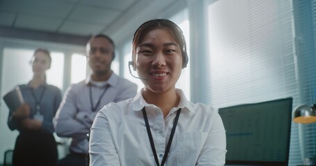 Call center office: Close up portrait of friendly Asian female technical customer support specialist wearing headset smiling, looking at camera. Team of hotline operators standing in the background.