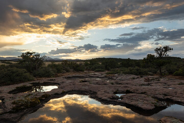 A recent rainstorm has left ephemeral pools on a sandstone ridge in the mountains of Southern Utah, USA. The setting sun casts beams of light across the clouds and the water reflects them. 