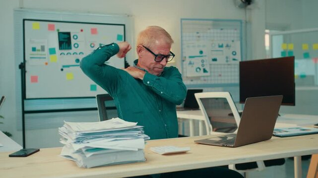 Office worker massaging his shoulder while surrounded by paperwork and a laptop. Signs of pain and stress are evident in the busy office environment, highlighting workplace challenges