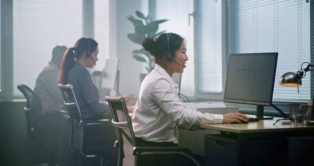 Diverse team of technical support operators in modern call center office. Asian female consultant talks on headset, works with data on computer, provides online technical support to customer over call