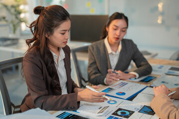 Asian businesswomen analyzing financial data and discussing company strategy during a productive meeting in modern office, using charts, calculators, and tablets