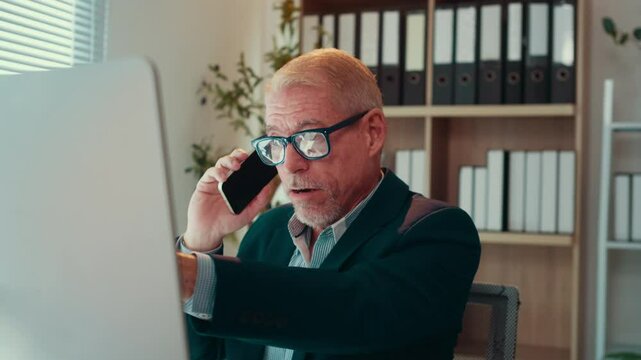 Businessman is sitting at his desk, talking on the phone and pointing at his computer screen. He is wearing a suit and glasses
