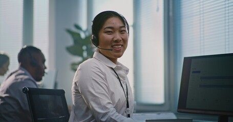 Asian female technical support operator wearing headset, sits in front of computer, looks at camera...