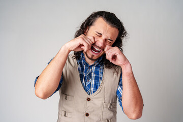 A man with curly hair wearing a plaid shirt expresses sadness while making a crying gesture in a plain background