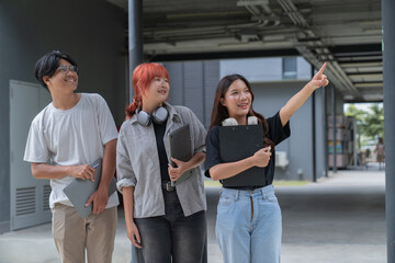 Obraz premium Three young students, holding laptops and clipboard, are standing together outdoors on a campus, with one student pointing towards something in the distance while engaging in a discussion