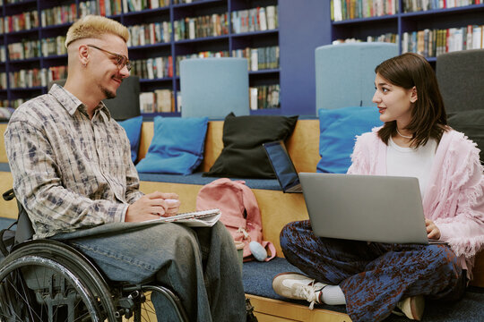 Two individuals engaging in a creative conversation in a modern library setting, with one person in a wheelchair and another holding a laptop, surrounded by shelves filled with books