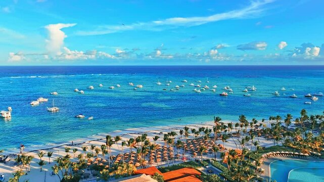 Luxury beach resort hotel, drone shot. Swimming pool and sun loungers, umbrellas under palm trees. Summer evening on the coast, tourist vacation background. Blue sea with white yachts.