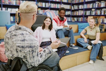 Group of diverse students sitting in modern library engaged in study session on tiered seating. One student in wheelchair, all focused and interacting, surrounded by shelves of books