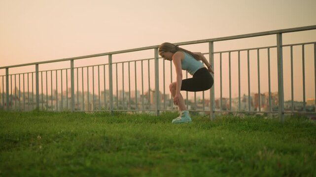 Young lady in roller skates holding iron railing while sitting on grassy ground, with beautiful evening cityscape in blurred background
