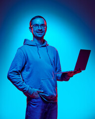 Young man, IT specialist looks confidently and smiling at camera holding laptop in neon light against gradient studio background. Concept of human emotions, beauty and fashion,